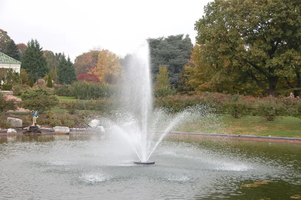 A fountain spraying in a pond at Hershey Gardens with autumn trees behind it