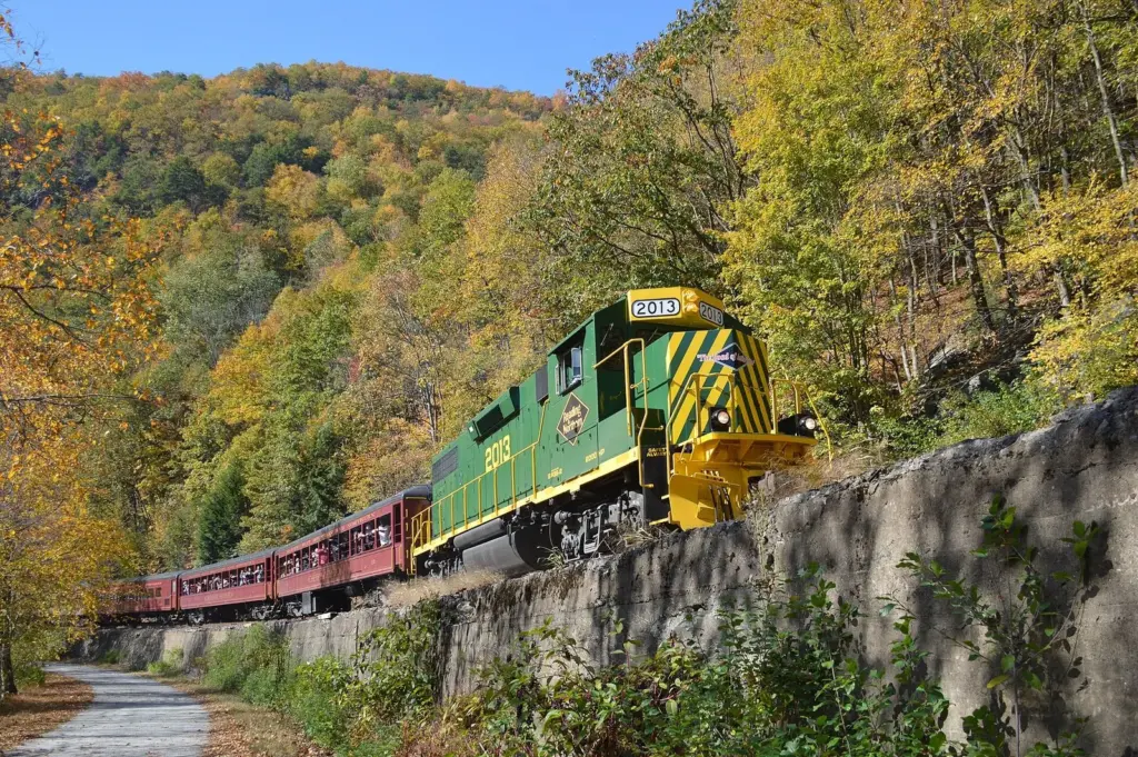 A green Reading and Northern diesel locomotive pulling red passenger cars below fall-colored hills along the Lehigh River