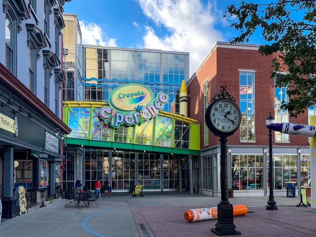 The glass entrance of the Crayola Experience with its colorful sign on a pedestrian plaza in Easton