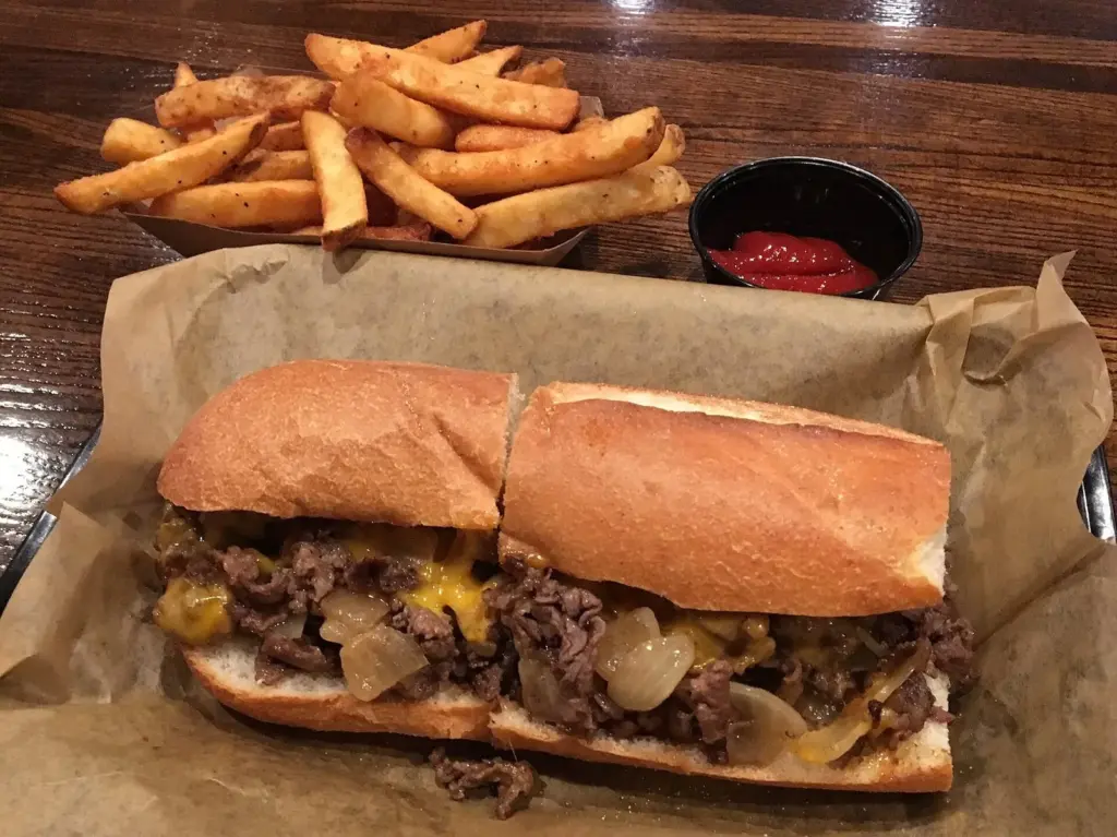 A cheesesteak with fried onions and melted cheese on a long roll, served with seasoned fries